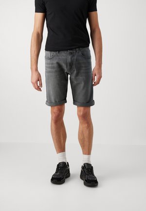 Homme portant un short en jean gris retroussé, un t-shirt noir, des chaussettes blanches et des baskets noires, debout devant un fond uni.