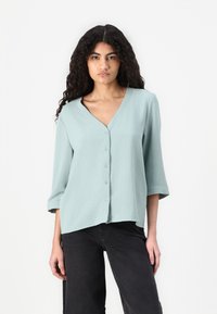 Blusa con scollo a V e bottoni in morbido tessuto verde mentolo testurizzato, con maniche a tre quarti, abbinata a pantaloni neri larghi.
