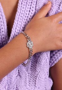 Zilveren kettingarmband met een gedraaide knoopontwerp als middelpunt, gedragen op een hand tegen een getextureerd lila gebreid kledingstuk.