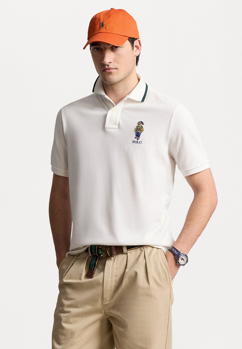 Polo shirt blanc avec des accents de col verts, logo brodé sur la poitrine. Porté avec un pantalon beige et une casquette couleur rouille. Montre au poignet.