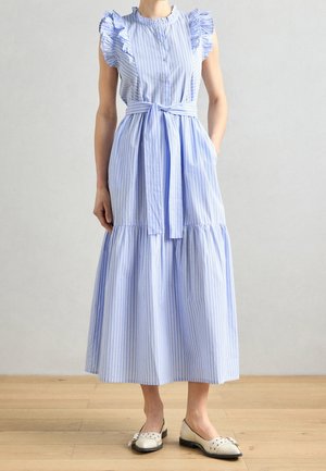 Frau trägt ärmelloses Midi-Kleid mit blau-weiß vertikalen Streifen, Rüschen an den Schultern, vorne geknöpft, Taillengürtel und weißen flachen Schuhen.