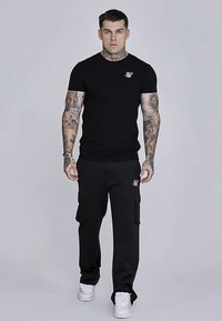 Svart t-shirt med kort ärm och en liten logga på bröstet, kombinerad med svarta cargobyxor med sidofickor och vita sneakers.