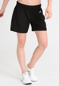 Nainen pukeutuneena mustiin Adidas-shortseihin ja valkoisiin Adidas-tennareihin, seisoo jalat ristissä vaaleaa taustaa vasten.