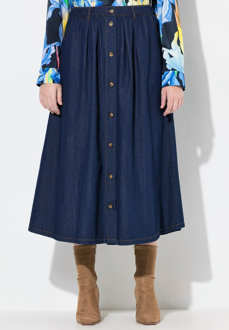 Jupe midi en denim avec un devant à boutons, dotée d'une taille élastique, de couleur bleu foncé et de détails de surpiqûre subtils. Des bottes ankle marron complètent le look.