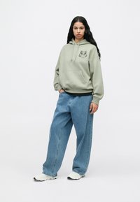 Fiatal nő hosszú sötét hajjal, világoszöld kapucnis felsőt viselve, lepke mintával, bő kék farmernadrágban és fehér tornacipőben áll egy fehér háttér előtt.