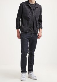 Musta kevyt takki, jossa on kaksi rintataskua, yhdistetty mustaan t-paitaan ja tummansinisiin farkkuihin. Valkoiset lenkkarit viimeistelevät asun.