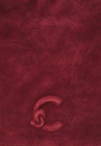 Produit en daim bordeaux avec une texture lisse, présentant un discret logo en relief dans la partie inférieure. Esthétique épurée et minimaliste.