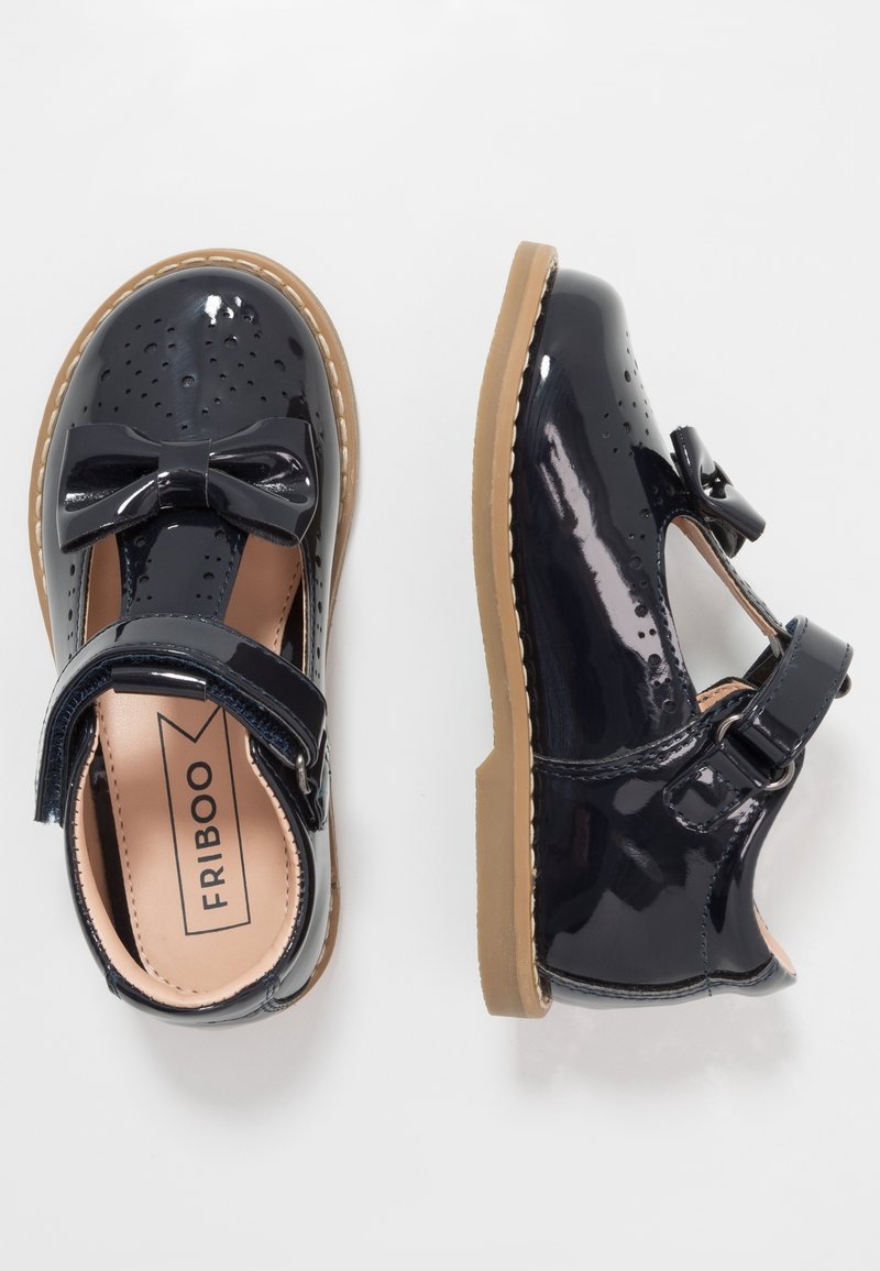 Paire de chaussures habillées noires vernies pour enfants avec nœuds, détails perforés, semelles beiges et brides à scratch, vue de dessus.