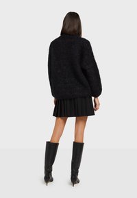 Oversized zwarte fuzzy sweater met lange mouwen, een ronde halslijn, gecombineerd met een zwarte plissé rok en kniehoge zwarte haklaarzen.