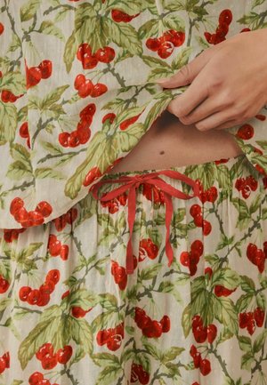 Hand hebt ein cherry-muster Oberteil an, das die gebundene Taille einer passenden, lockeren Hose mit Kordelzug auf bloßer Haut zeigt.