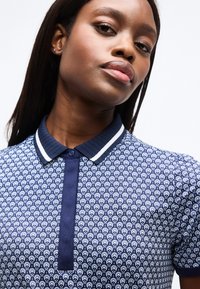 Νεαρή γυναίκα με μακριά σκούρα μαλλιά που φοράει μπλε ναυτική πόλο μπλούζα με λευκή λεπτομέρεια στο γιακά, σε λευκό φόντο.