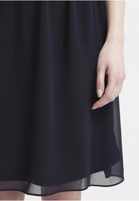 Abito in chiffon blu scuro con un design fluido, caratterizzato da una texture a strati e un orlo liscio. Una mano parzialmente visibile in basso.