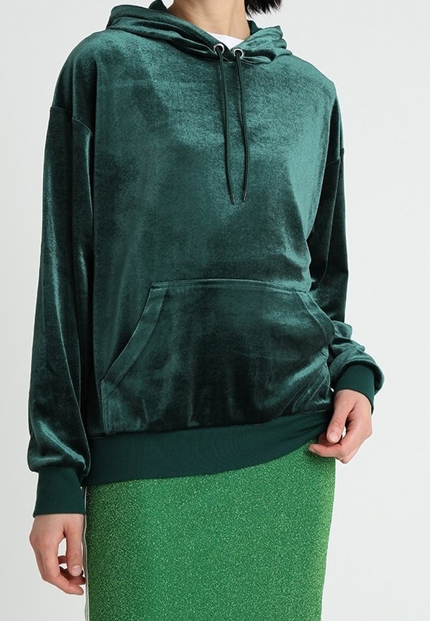 Personne portant un sweat à capuche en velours vert foncé avec une poche avant et des cordons, associé à une jupe texturée vert vif.