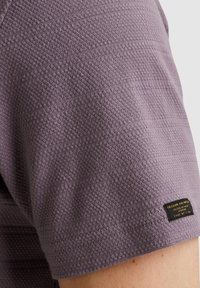 Camisa corta de manga corta texturizada en color púrpura con un patrón tejido y una pequeña etiqueta negra cerca del dobladillo con detalles de la marca.