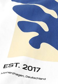 Blaues abstraktes Shape auf beigem Quadrat mit schwarzem Text "EST. 2017 Meinerzhagen, Deutschland" auf weißem Stoffhintergrund.