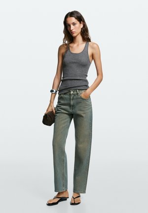 Junge Frau trägt ein graues Tanktop, ausgewaschene Jeans mit hoher Taille, schwarze Sandalen, hält eine kleine braune Clutch und trägt ein silbernes Armband.