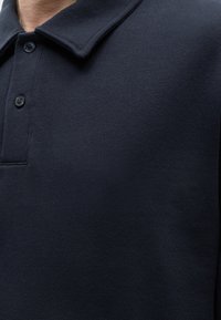 Κοντινή λήψη ενός ατόμου που φορά μπλε σκούρο μπλουζάκι πόλο με γιακά και δύο ορατά κουμπιά.