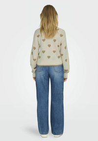Beiger Pullover mit hellbraunen Herzmustern, gerippten Bündchen und Saum. Kombiniert mit lockeren blauen Jeans und weißen Schuhen. Rückansicht.