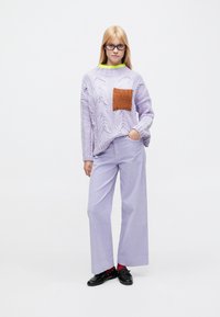 Suéter grande de punto lila con un patrón texturizado y un bolsillo parche naranja, combinado con pantalones lila a rayas de pierna ancha. Se ven zapatos negros.
