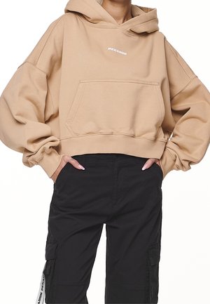 Beige, kurz geschnittenes Hoodie mit Fronttasche und kleinem "PEGADOR"-Logo, kombiniert mit schwarzer Cargohose mit Seitentaschen und weißen Riemendetails.