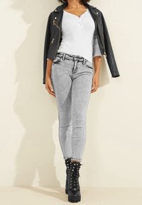 Donna che indossa jeans skinny grigi chiari, una maglietta bianca a maniche lunghe, una giacca di pelle nera sulle spalle e stivali da combattimento neri stringati.