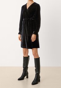 Svart sammet omslagklänning med långa puffärmar och midjeband. Bärs med knähöga svarta läderstövlar med klack.