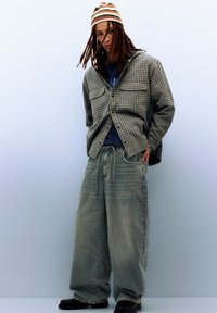 Casual outfit met een geruite jas, blauw T-shirt, losse lichtgewassen spijkerbroek en een gestreepte beanie. Zwarte instapschoenen.