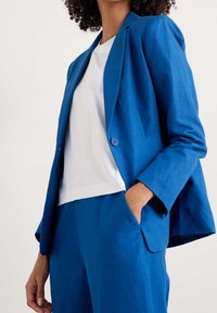 Blå linn blazer med avslappnad passform, enkel knäppning och lätt uppvikta ärmar, i kombination med en vit t-shirt och matchande byxor.