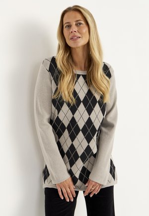 Kvinde med langt blondt hår iført en beige og sort argyle sweater og sorte bukser, der står op ad en hvid væg.