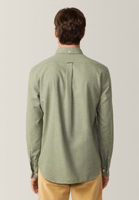 Camiseta verde de manga larga con tejido texturizado, cuello puntiagudo, puños abotonados y un pliegue en la parte trasera para un mejor ajuste. Lleva puesta con pantalones de color claro.