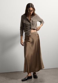 Cardigan de lana marrón con dos bolsillos frontales, combinado con una falda larga y fluida de satén marrón y zapatos planos negros sobre un fondo neutro.