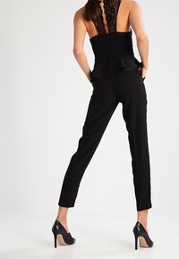 Mono negro con parte superior de encaje en la espalda, corsé ajustado, detalle de peplum y piernas ajustadas. Llevado con tacones altos negros, mostrando una silueta elegante.