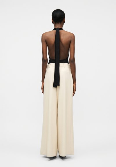 Femme de dos portant un haut dos nu noir à col halter avec une longue attache et un pantalon beige taille haute à jambes larges sur fond uni.