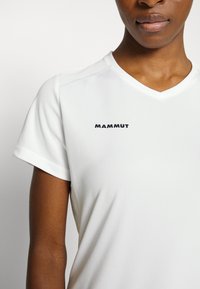 Primer plano de una persona que lleva una camiseta blanca de cuello en V con el logo "MAMMUT" en el pecho contra un fondo liso.