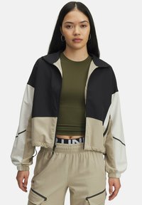 Jeune femme portant une veste zippée colorée en noir, beige et blanc sur un top court olive vert et un pantalon cargo beige.