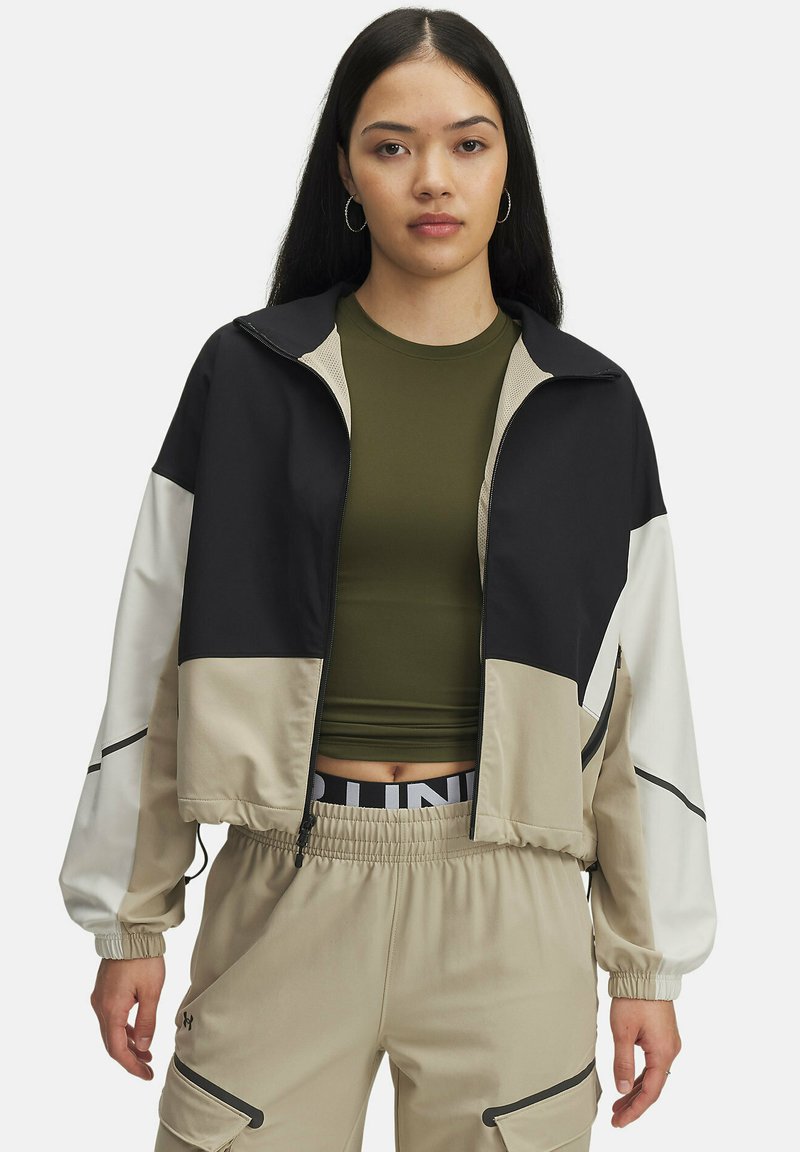 Jeune femme portant une veste zippée colorée en noir, beige et blanc sur un top court olive vert et un pantalon cargo beige.
