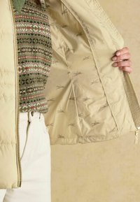 Béžová prešívaná bunda s prešívaným povrchom a zlatým zipsom. Interiér má vzorovanú podšívku s malými motívmi koní. Spárovaná so zeleným svetrom.