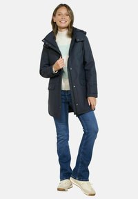 Parka imperméable bleu marine avec une capuche amovible, des poches à rabat boutonnées et une fermeture éclair. Portée par-dessus un pull léger et un jean.