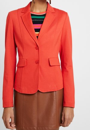 Vrouw draagt een nauwsluitende fel koraalrode blazer met klepzakken over een gestreepte meerkleurige top en een bruine leren rok.