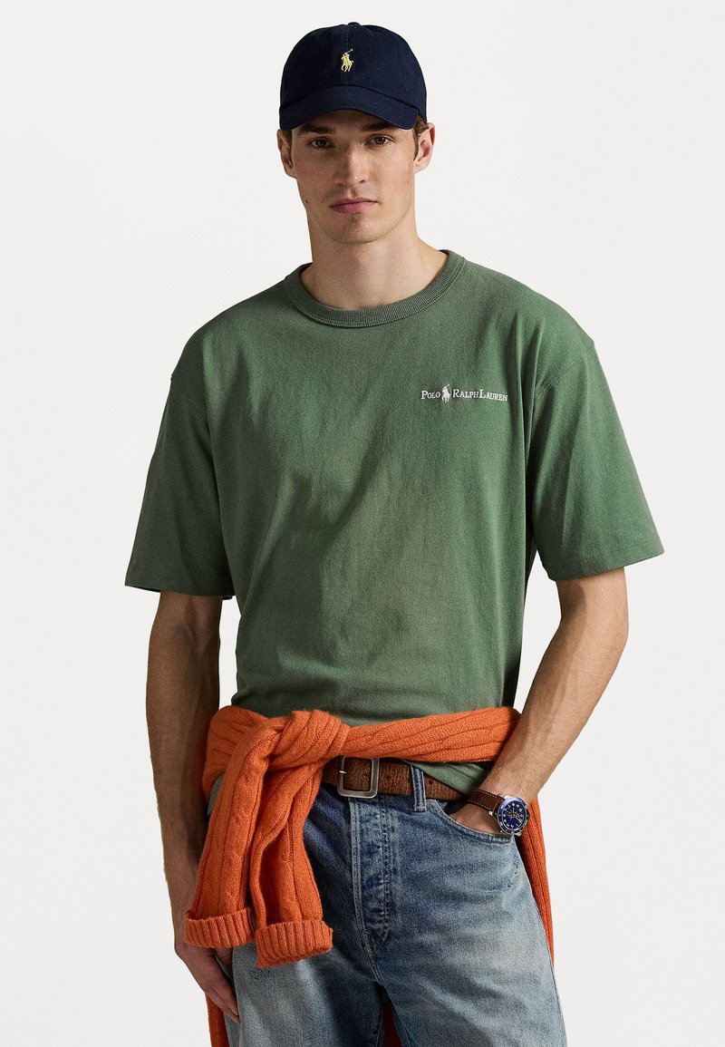 Green cotton t-shirt with a crew neck and short sleeves, paired with rolled-up orange knit sweater tied around the waist. Wearing a navy cap.