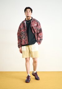 Rød og svart mønstret jakke, svart skjorte, beige shorts med hvite detaljer, mørke joggesko. Mann som står mot en nøytral bakgrunn.