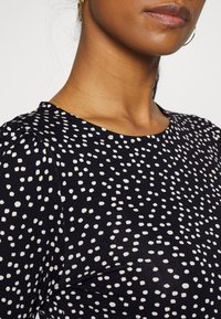 Tecido preto com padrão de bolinhas brancas, com decote redondo e detalhe de ombro levemente franzido, apresentando textura suave sem adornos.