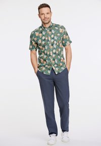 Camicia verde a maniche corte con motivo floreale in tonalità di arancione e blu, abbinata a pantaloni blu scuro e sneakers bianche.