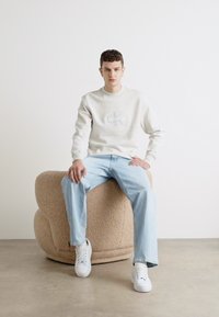 Sudadera gris claro con el logo de CK, combinada con jeans azul claro. Modelo sentado en una silla beige redondeada y texturizada. Zapatillas blancas completan el look.