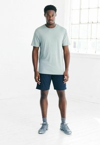 Hellgrünes Sport-T-Shirt, marineblaue Shorts, graue Sportschuhe, steht auf einem weißen Boden in einem gut beleuchteten Raum, die Hände entspannt an den Seiten.