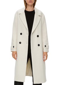 Dubbelknäppt kappa i texturerat vitt tygl med svarta knappar, vida slag och sidospjäll för rörelsefrihet.