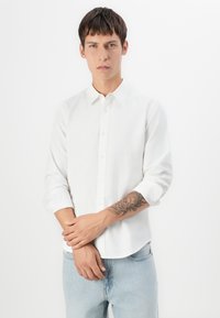 Joven con cabello corto castaño que lleva una camisa blanca abotonada y jeans azul claro, mostrando un tatuaje en su antebrazo izquierdo.
