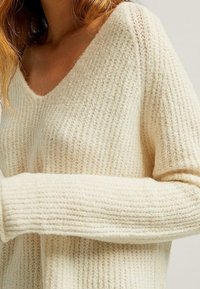 Femme portant un pull en maille côtelée de couleur crème, avec un col en V et des manches longues, vu des épaules à la taille.