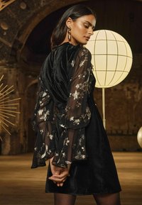 Rochie din catifea neagră cu mâneci transparente negre, decorate cu modele de stele argintii. Silueta este ajustată, cu mâneci evazate și volane.