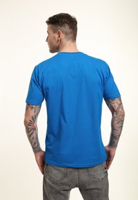 Blue short-sleeved T-shirt made of cotton, featuring a round neckline. Back view shows smooth texture and a relaxed fit. Grey jeans visible below.